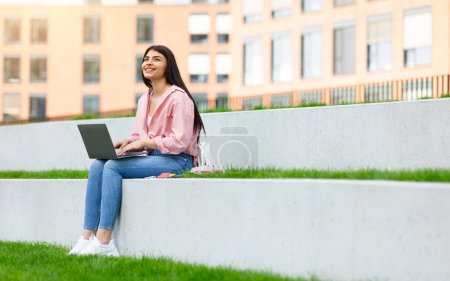 Lächelndes junges Mädchen genießt einen sonnigen Tag, während sie draußen an ihrem Laptop vor dem Hintergrund von Gebäuden arbeitet