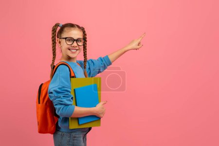 Foto de Estudiante joven con gafas y mochila apuntando hacia un lado sobre un fondo rosa, espacio para copiar - Imagen libre de derechos