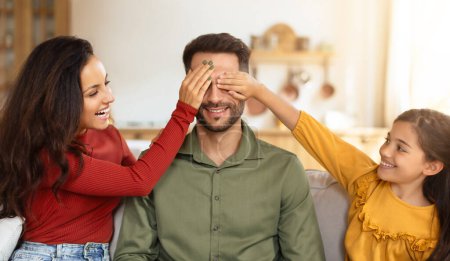 Photo for A fun, playful moment with a mother and daughter covering her husbands father eyes as they enjoy family time on the sofa - Royalty Free Image