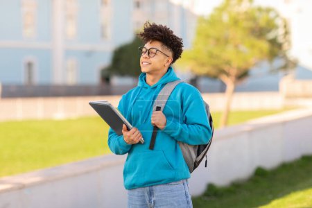 Téléchargez les photos : Un jeune étudiant cher tenant une tablette et portant un sac à dos marche dehors dans un cadre de campus avec un look serein - en image libre de droit