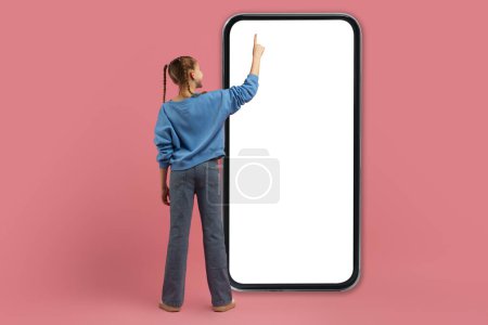 Téléchargez les photos : Une jeune fille en tenue décontractée tend la main vers un écran géant vierge de smartphone sur un fond rose - en image libre de droit