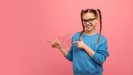 Photo for Smiling young girl with glasses points sideways, suggesting choice or direction, against pink background - Royalty Free Image