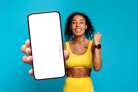 Foto de Alegre entusiasta del fitness dama afroamericana que presenta una pantalla de teléfono inteligente en blanco con una sonrisa brillante sobre un fondo azul - Imagen libre de derechos