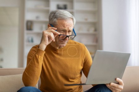Konzentrierter älterer Mann, der einen Laptop für Arbeit oder Freizeit in seinem Wohnhaus benutzt und die Brille anpasst