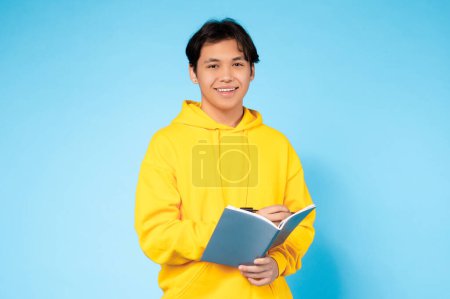 Téléchargez les photos : Un asiatique détendu dans un sweat-shirt jaune confortable est absorbé dans la lecture d'un livre avec un fond bleu - en image libre de droit