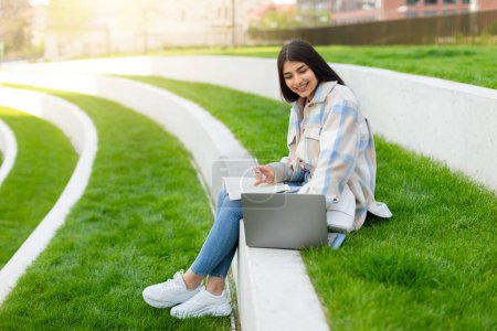 A young girl sits on a curved bench in an outdoor setting, working on her laptop with a notebook besides her
