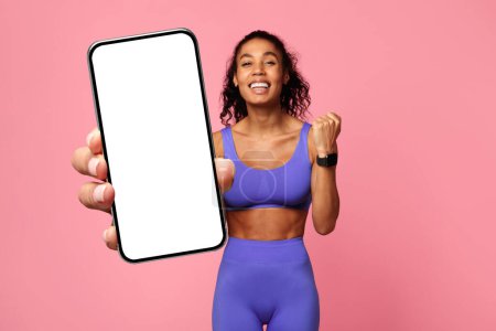 Téléchargez les photos : Femme afro-américaine énergique dans un vêtement de sport bleu montrant un smartphone avec un écran vide, exprimant l'excitation sur un fond rose - en image libre de droit