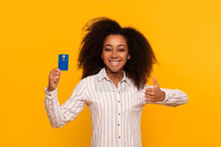 Téléchargez les photos : Sourire jeune femme afro-américaine présentant carte de crédit et signe d'approbation sur un fond jaune - en image libre de droit