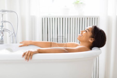 Téléchargez les photos : Jeune femme afro-américaine sereine se relaxant dans une baignoire remplie, les yeux fermés dans une salle de bain immaculée - en image libre de droit