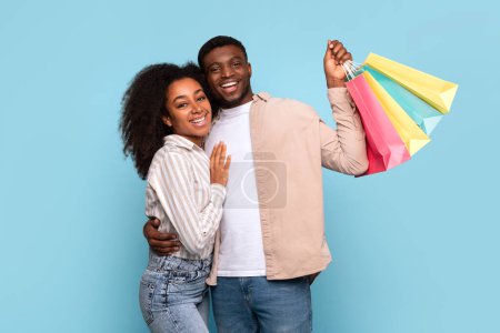 Ein fröhliches junges afroamerikanisches Paar umarmt sich, als der Mann bunte Einkaufstüten in der Hand hält, die auf einen erfolgreichen Einkaufsbummel hindeuten.