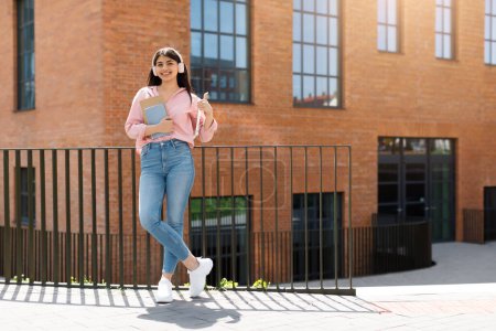 Téléchargez les photos : Jeune fille étudiante marche dans le but tout en tenant un smartphone, avec les bâtiments du campus derrière elle - en image libre de droit