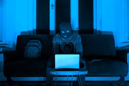 A mysterious figure burglar sits before a glowing laptop screen, creating a striking contrast in a dark, moody room with a blue overtone, invoking curiosity and intrigue