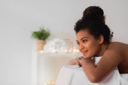 A young african american woman with curly hair smiles gently while wrapped cozily in a white blanket, with soft lighting in the background