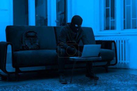 Foto de Un ladrón cibernético enfocado en un pasamontañas se sienta trabajando en una computadora portátil en una habitación con poca luz, emanando una sensación de peligro y secreto. - Imagen libre de derechos