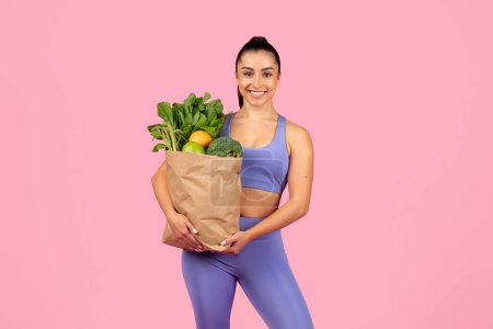 Photo for Smiling woman in fitness attire holds a paper bag stuffed with fresh, healthy groceries - Royalty Free Image