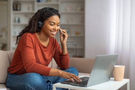 Foto de Una mujer negra alegre con auriculares está participando en una conversación en línea utilizando un ordenador portátil en un sofá acogedor, mujer afroamericana haciendo videollamada en casa - Imagen libre de derechos