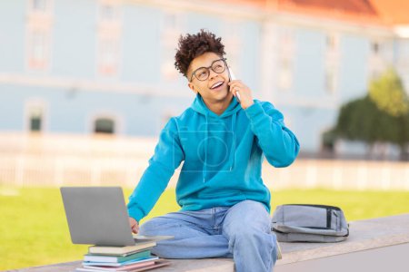 Foto de Sonriente brasileño chico estudiante habla en el teléfono móvil mientras trabaja con un ordenador portátil fuera en un entorno del campus con mochila y libros - Imagen libre de derechos