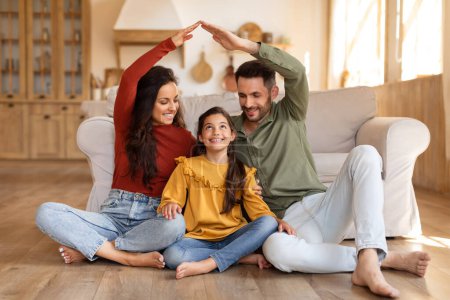 Lächelnde Familie schafft eine herzerwärmende Szene, indem sie mit ihren Händen eine Hausform gestaltet, während sie auf dem Boden sitzt