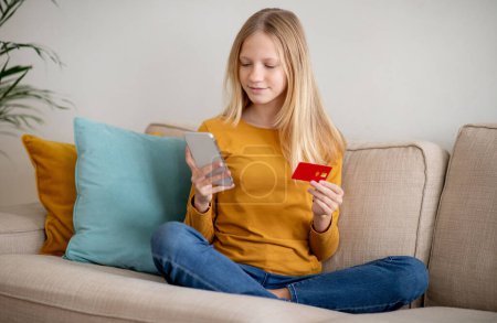 Chica adolescente sentada cómodamente en un sofá, sosteniendo su tarjeta de crédito y navegando en el teléfono inteligente, sonriendo adolescente compras en línea, utilizando la aplicación moderna para las compras