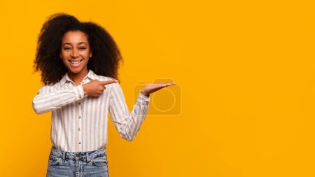 Téléchargez les photos : Une jeune femme afro-américaine joyeuse aux cheveux bouclés, pointant et présentant quelque chose sur son côté sur un fond jaune vif - en image libre de droit