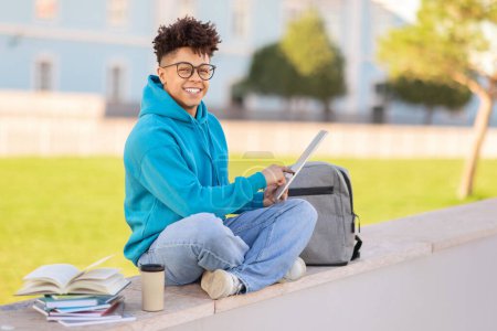 Photo for A brazilian guy student in casual attire is working on a tablet while sitting on a ledge with textbooks and a backpack beside - Royalty Free Image