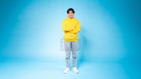 A cheerful young Asian guy stands confidently with arms crossed, wearing a bright yellow hoodie against a vivid blue background