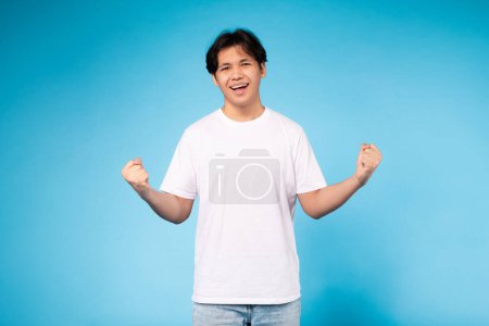 An enthusiastic young Asian guy throwing his hands up in a celebratory pose on a blue backdrop, celebrating success