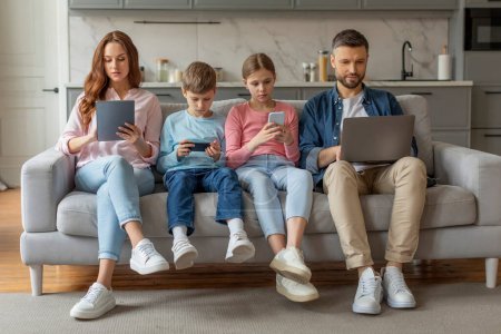 A family of four engrossed in digital devices while sitting together on a sofa in a cozy home setting