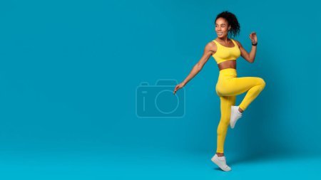 Dynamic shot of a vivacious African American woman in yellow fitness clothing captured in mid-run on a blue background