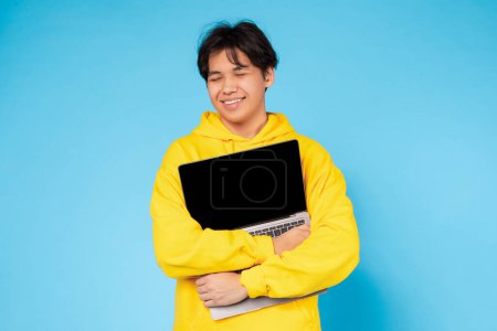 Téléchargez les photos : Tenu par un adolescent asiatique de contenu, un ordinateur portable avec un écran noir est prêt pour un contenu personnalisé, sur fond bleu - en image libre de droit
