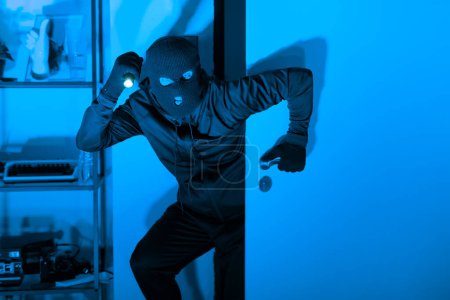 Téléchargez les photos : Un criminel sournois avec une lampe de poche et un masque de ski capturés en mouvement lors d'un cambriolage dans une pièce éclairée par une lumière bleue étrange - en image libre de droit