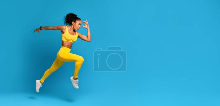 Energetic young African American woman wearing yellow sports clothing captured in a dynamic mid-air jump on a blue background