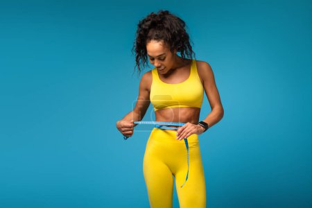 Afroamericana consciente de la aptitud vestida de gimnasia amarilla midiendo su cintura con una cinta métrica sobre un fondo azul