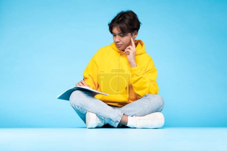 Foto de Joven adolescente asiático en suéter amarillo meditando mientras está sentado con las piernas cruzadas con un libro abierto sobre fondo azul - Imagen libre de derechos