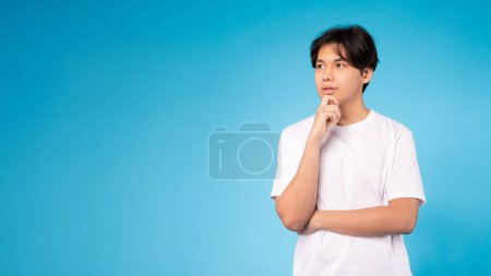 Joven adolescente asiático meditando con su mano sobre su barbilla y mirando hacia arriba, de pie sobre un fondo azul