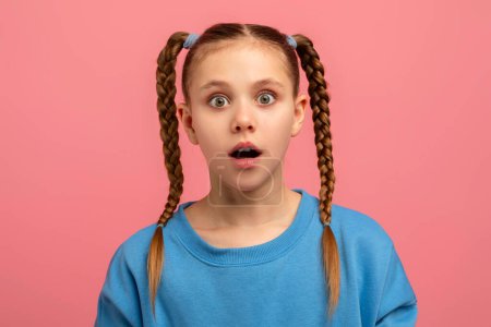 Shocked girl with braided hair and wide eyes stands against a pink studio background, showing surprise
