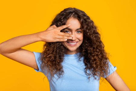 Téléchargez les photos : Femme souriante couvrant ludique son visage avec sa main et regardant à travers les doigts sur fond jaune - en image libre de droit