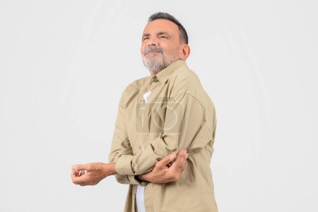 Ein älterer Herr im beigen Hemd reibt sich den Ellbogen, posiert auf weißem Hintergrund und hat Schmerzen im Arm