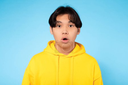Primer plano de un joven asiático en una sudadera con capucha amarilla, expresión facial de sorpresa con un fondo azul