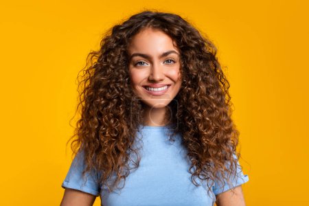 Mujer joven accesible con una sonrisa cálida y el pelo rizado posando sobre un fondo amarillo, primer plano