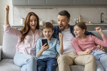 Joyful family raises their fists triumphantly while looking at a smartphone on a sofa at home, domestic entertainment