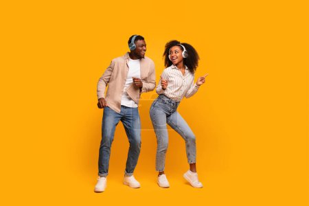 Téléchargez les photos : Couple afro-américain dansant et appréciant la musique portant des écouteurs sans fil sur un fond de studio jaune - en image libre de droit