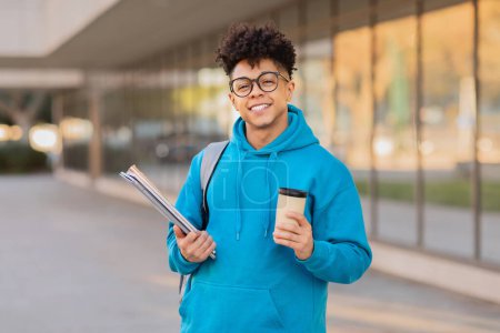 Fröhlicher junger brasilianischer Student mit Büchern und Kaffeetasse, mit blauem Kapuzenpulli und Brille im Freien