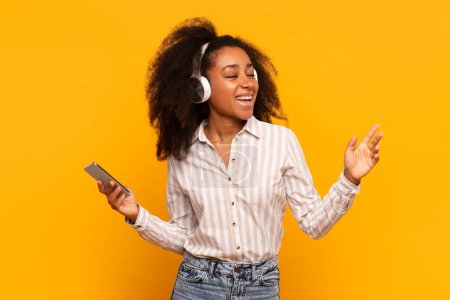 Photo for An exuberant African American woman dances while listening to music, eyes closed in contentment against a vibrant yellow background - Royalty Free Image