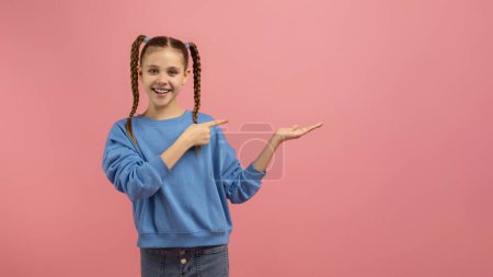 Photo for A smiling girl with braids presenting something with a hand gesture, wearing a blue sweater on a pink background - Royalty Free Image