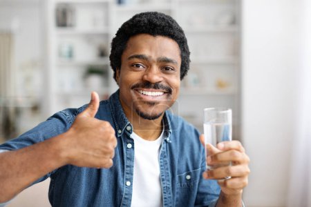 A cheerful black man in denim shirt holding clear glass of water and gesturing thumb up, african american male showing sign of approval, copy space