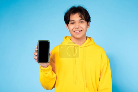 Adolescent Asian guy in yellow sweatshirt confidently displaying a blank smartphone screen, ideal for mockups, against a blue studio backdrop