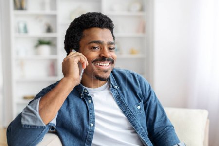 Téléchargez les photos : Un jeune homme noir joyeux assis confortablement tout en parlant sur son téléphone portable à la maison. - en image libre de droit