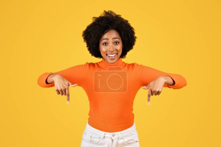 Foto de Mujer joven radiante con el pelo rizado sonriendo ampliamente y apuntando hacia abajo con ambas manos, usando un cuello alto naranja brillante y pantalones blancos sobre un fondo amarillo llamativo - Imagen libre de derechos