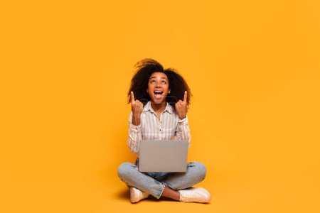 Téléchargez les photos : Joyeuse jeune femme afro-américaine aux cheveux bouclés s'assoit jambes croisées sur le sol, à l'aide d'un ordinateur portable et pointant vers le haut à l'espace de copie - en image libre de droit